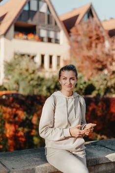 Young fashionable teenage girl with smartphone in park in autumn sitting at smiling. Trendy young woman in fall in park texting. Retouched, vibrant colors. Beautiful blonde teenage girl wearing casual modern autumn outfit sitting in park in autumn. Retouched, vibrant colors, brownish tones.