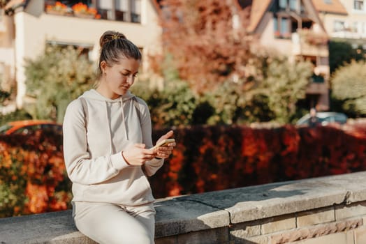 Young fashionable teenage girl with smartphone in park in autumn sitting at smiling. Trendy young woman in fall in park texting. Retouched, vibrant colors. Beautiful blonde teenage girl wearing casual modern autumn outfit sitting in park in autumn. Retouched, vibrant colors, brownish tones.