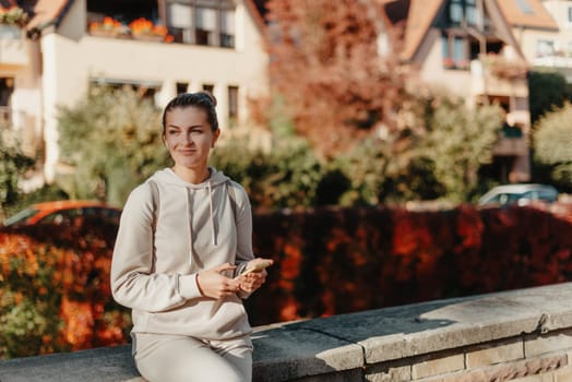 Young fashionable teenage girl with smartphone in park in autumn sitting at smiling. Trendy young woman in fall in park texting. Retouched, vibrant colors. Beautiful blonde teenage girl wearing casual modern autumn outfit sitting in park in autumn. Retouched, vibrant colors, brownish tones.