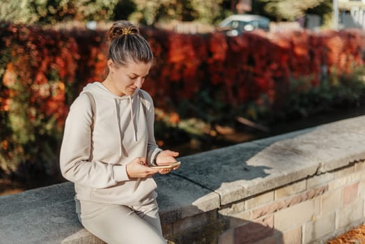 Young fashionable teenage girl with smartphone in park in autumn sitting at smiling. Trendy young woman in fall in park texting. Retouched, vibrant colors. Beautiful blonde teenage girl wearing casual modern autumn outfit sitting in park in autumn. Retouched, vibrant colors, brownish tones.