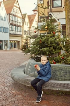 Winter Wonderland Delight: 8-Year-Old Boy with Christmas Decor by Vintage Fountain. Experience the magic of winter joy with this enchanting image featuring a beautiful 8-year-old boy sitting on a stone bench against the backdrop of a festively decorated vintage fountain. The charming scene unfolds in a quaint German town with half-timbered houses adorned with holiday lights, creating a picturesque winter setting. The boy holds a red Christmas ornament, adding a touch of festive warmth to the vintage-toned photograph.
