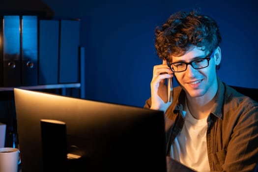 Young businessman calling on smartphone to customer or coworker, looking at computer screen of creative presentation surround work desk at neon modern office. Concept of new next project. Gusher.