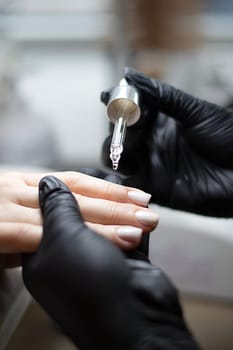 Beauty concept. A manicurist in black latex gloves makes a hygienic manicure and paints the client's nails with gel polish and drips oil on the cuticles in a beauty salon. Close-up. Vertical.