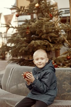 Winter Wonderland Delight: 8-Year-Old Boy with Christmas Decor by Vintage Fountain. Experience the magic of winter joy with this enchanting image featuring a beautiful 8-year-old boy sitting on a stone bench against the backdrop of a festively decorated vintage fountain. The charming scene unfolds in a quaint German town with half-timbered houses adorned with holiday lights, creating a picturesque winter setting. The boy holds a red Christmas ornament, adding a touch of festive warmth to the vintage-toned photograph.