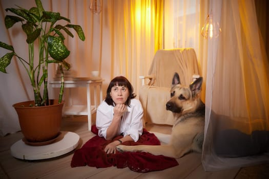 Adult mature woman with big shepherd dog in white shirt. Room with girl and calm cozy evening atmosphere with transparent curtains and soft warm light of lamps. Concept of love for animals, pets