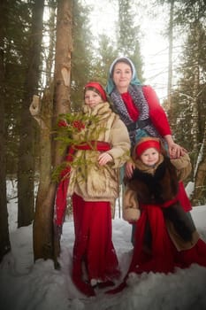 Family with mother, teenage girl, and little daughter dressed in stylized medieval peasant clothing in winter forest. Woman and her daughters pose for fairytale photoshoot in nature on a cold day