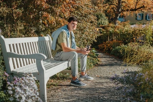 a teenager sits on a bench in the park drinks coffee from a thermo mug and looks into a phone. Portrait of handsome cheerful guy sitting on bench fresh air using device browsing media smm drinking latte urban outside outdoor.