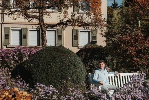 a teenager sits on a bench in the park drinks coffee from a thermo mug and looks into a phone. Portrait of handsome cheerful guy sitting on bench fresh air using device browsing media smm drinking latte urban outside outdoor.