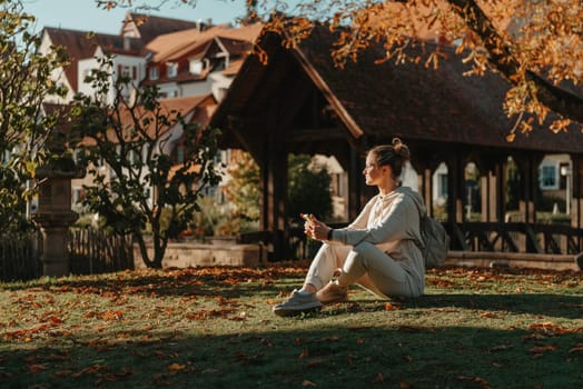 Young fashionable teenage girl with smartphone in park in autumn sitting at smiling. Trendy young woman in fall in park texting. Retouched, vibrant colors. Beautiful blonde teenage girl wearing casual modern autumn outfit sitting in park in autumn. Retouched, vibrant colors, brownish tones.