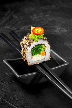 Vegetarian sushi roll with light filling of cream cheese, carrot, hiyashi wakame, lettuce and cucumber adorned with sesame seeds and chili, perfectly balanced on chopsticks over soy sauce dish