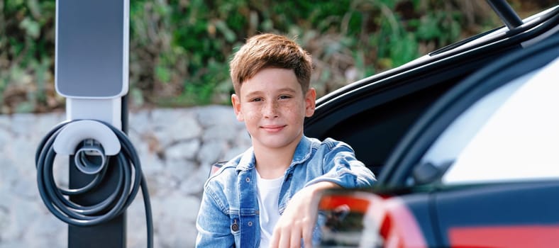 Little boy sitting on car trunk while recharging eco-friendly electric car from EV charging station. EV car road trip travel with alternative vehicle powered sustainable energy. Panorama Perpetual