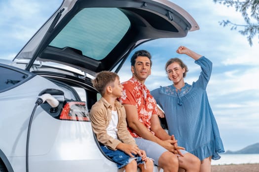 Family vacation trip traveling by the beach with electric car, lovely family sit on the trunk, charging EV car battery with green and sustainable energy. Family travel and eco-friendly car. Perpetual