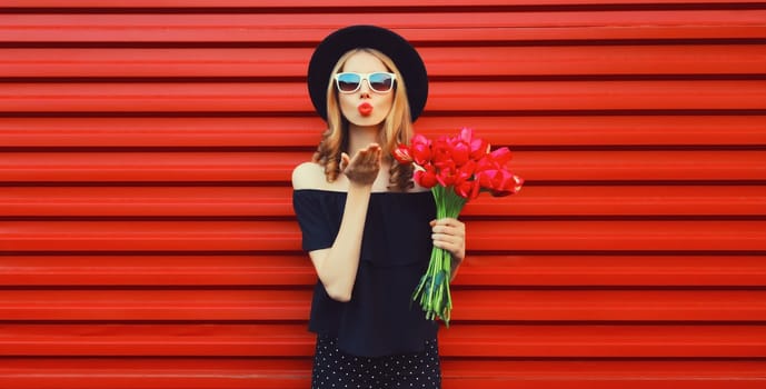 Portrait of beautiful stylish woman with bouquet of red rose flowers blowing kiss in black round hat
