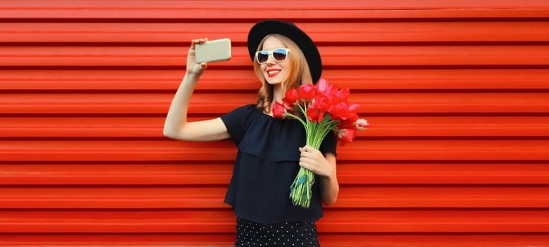 Beautiful young woman taking selfie with smartphone holding bouquet of red rose flowers in black round hat