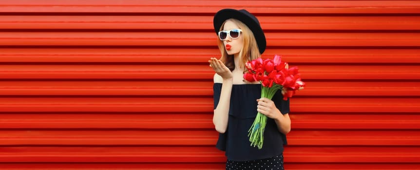 Portrait of beautiful stylish woman with bouquet of red rose flowers blowing kiss in black round hat
