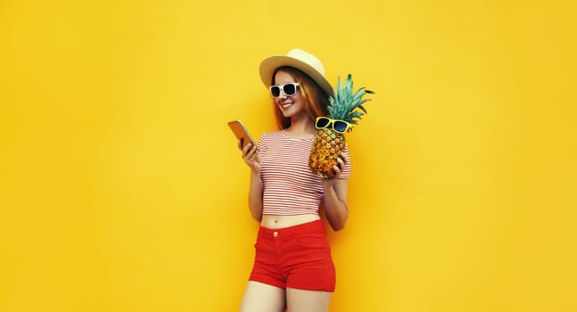 Summer portrait of traveler happy young woman with mobile phone holding fresh pineapple fruit looking at device in casual straw hat, sunglasses on bright yellow studio background