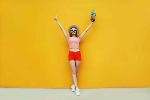 Summer bright image of cheerful young woman full length having fun holding pineapple, emotional girl raising her hands up with fruit on colorful yellow studio background