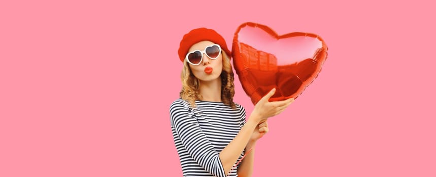 Sweet portrait of beautiful happy young woman with red heart shaped balloon blows kiss in french beret hat on pink studio background