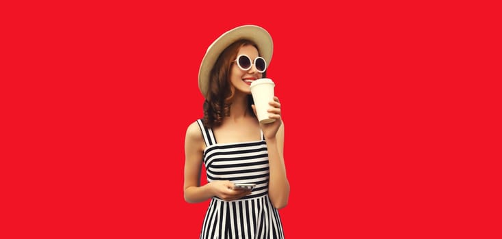 Happy smiling young woman drinking fresh juice or coffee and looking away wearing summer straw hat, striped dress on red studio background
