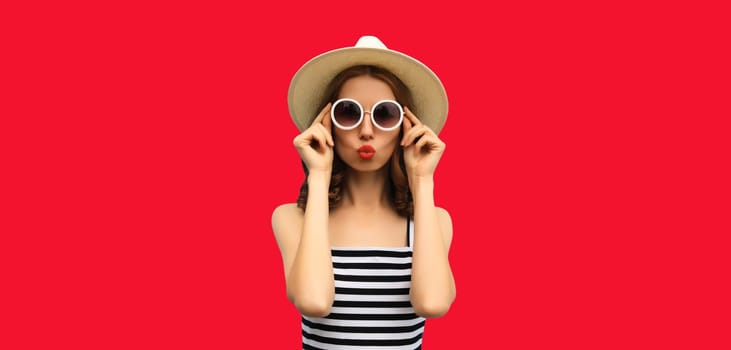 Beautiful young woman posing blowing kiss in summer straw hat on red studio background