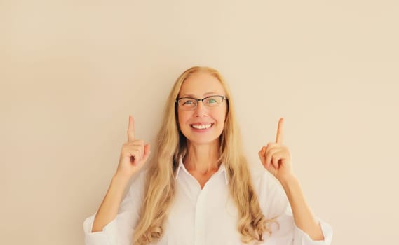 Happy exciting smiling middle aged woman in eyeglasses pointing her fingers up and looking at camera