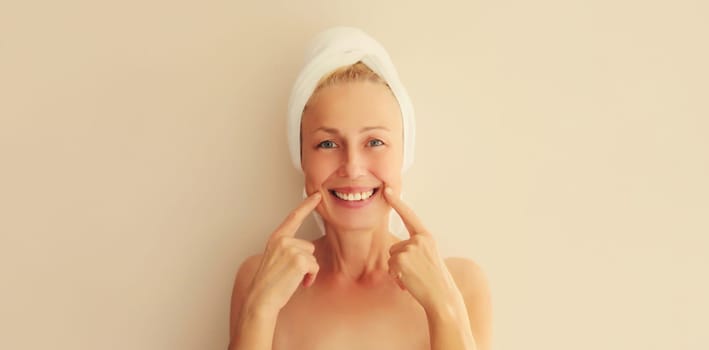 Happy smiling middle-aged woman points to her white clean teeth drying her wet hair with wrapped towel on head after shower in morning at home