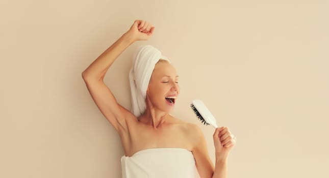 Happy cheerful woman having fun sings with hair comb and laughing after shower drying wet hair with bath towel on head in morning at home