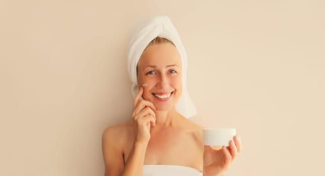 Natural beauty portrait of happy middle-aged woman touches her clean skin applying face cream with bath towel on head after shower in morning