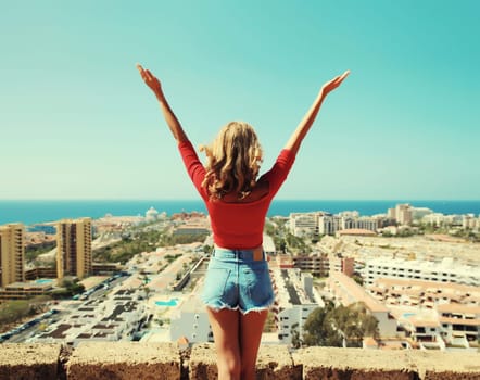 Summer vacation, beautiful happy young woman tourist raising her hands up on top of the mountain looking around the city in Tenerife, Canary Islands, Spain. Aerial top view.