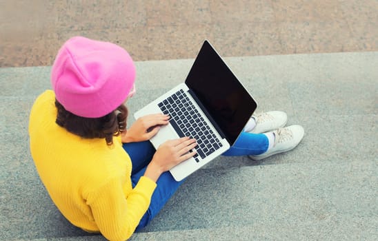 Stylish modern young woman working with laptop in colorful clothes in the city