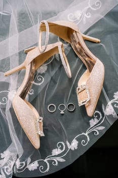 Wedding rings lie on an embroidered veil near the bride golden high-heeled shoes. High quality photo