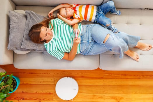 Mother and Child Relaxing on Couch with Smartphone and Robot Vacuum.