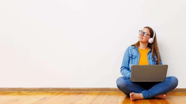 A relaxed young female in casual wear sitting cross-legged on the floor, listening to music with headphones and using a laptop.