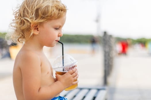 A young child with curly hair sipping a cold beverage through a straw from a plastic cup while sitting outside.