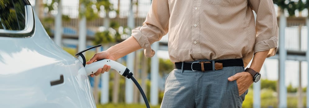 Young man recharge electric car's battery from charging station in city commercial parking lot. Rechargeable EV car for sustainable environmental friendly urban travel. Panorama Expedient