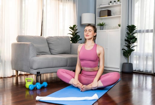 Flexible and dexterity woman in sportswear doing yoga position in meditation posture on exercising mat at home. Healthy gaiety home yoga lifestyle with peaceful mind and serenity.