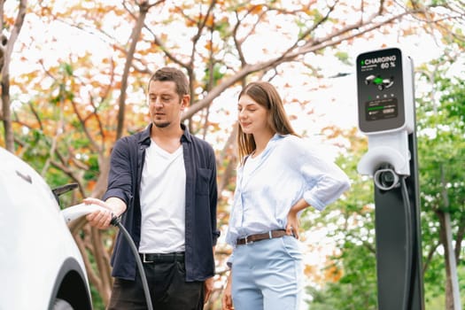 Lovely young couple recharging battery for electric car during autumnal road trip travel EV car in autumnal forest. Eco friendly travel on vacation during autumn. Exalt