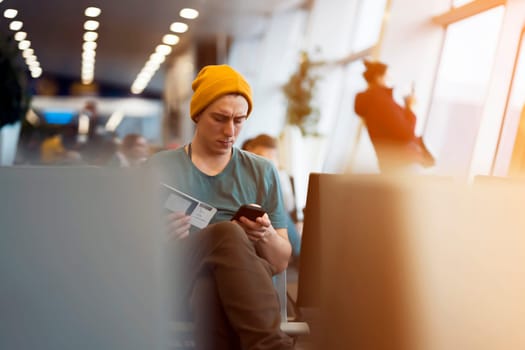A young man sits at the airport and checks in for the next flight via an app on his phone, waits for the plane and works while traveling.