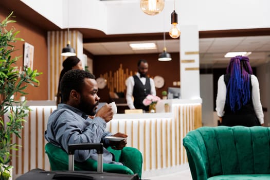 African American businessman drinking coffee while waiting for check-in procedure in hotel lobby. Tired black man traveler holding cup of espresso resting after long flight, waiting for room