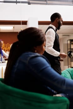 African American hotel staff and guest in lobby. Woman traveler waiting for check-in at reception area, friendly smiling bellboy helping guests with luggage. Hospitality and tourism Industry