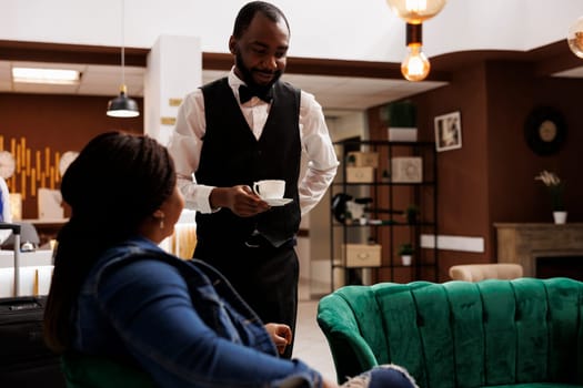 Pleasant smiling hotel worker offering free coffee to tired guest waiting for check-in process in lobby, waiter serving cappuccino to tourist arriving at resort. Customer service in hospitality