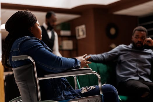 Disabled hotel guest sitting in lobby, African American woman in wheelchair at sitting area in foyer, acomodations for travelers with reduced mobility. Disability access in hospitality industry