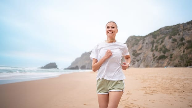 Smiling young woman jogging on beach along sea waves. Fit female jogger in white t-shirt and shorts running on sandy beach. Woman runner represents healthy lifestyle.