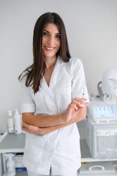 Portrait of smiling female cosmetologist holding Botox injection in clinic. Female dermatologist wearing white medical uniform holding syringe and looking at camera. Brunette female doctor.