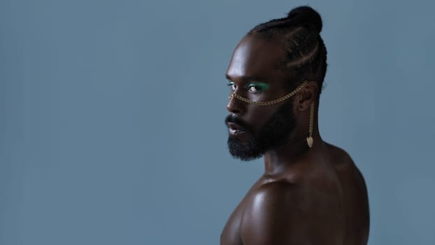 Side view of bearded African American gay man with light makeup and gold accessory on face. Senior shirtlesss adult homosexual male looking at camera with mockup space on blue background.