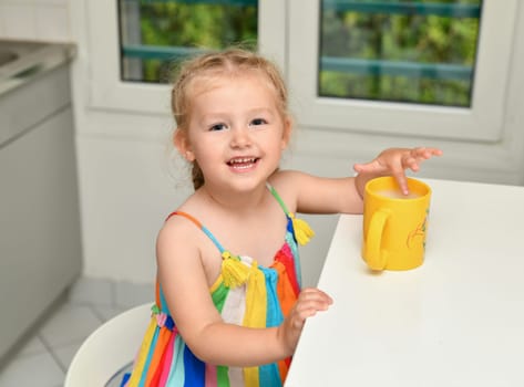 A girl in a rainbow dress sits in the kitchen and drinks cocoa with milk