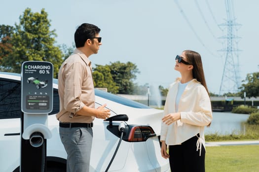 Young couple recharge EV car battery at charging station connected to power grid tower electrical industrial facility as electrical industry for eco friendly vehicle utilization. Expedient