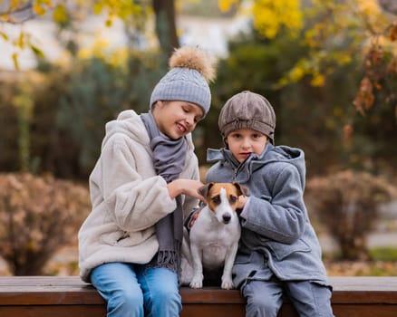 Brother and sister sit in an embrace with a dog on a bench for a walk in the autumn park. Boy, girl and jack russell terrier