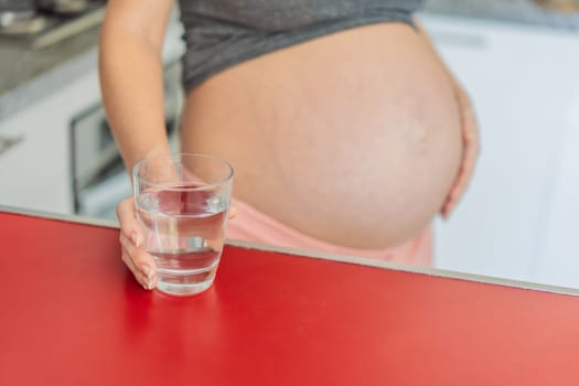 Embracing the vital benefits of water during pregnancy, a pregnant woman stands in the kitchen with a glass, highlighting hydration's crucial role in maternal well-being.