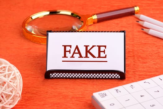 FAKE word written on a white business card on a stand on an orange background, next to a magnifying glass, a calculator and pencils